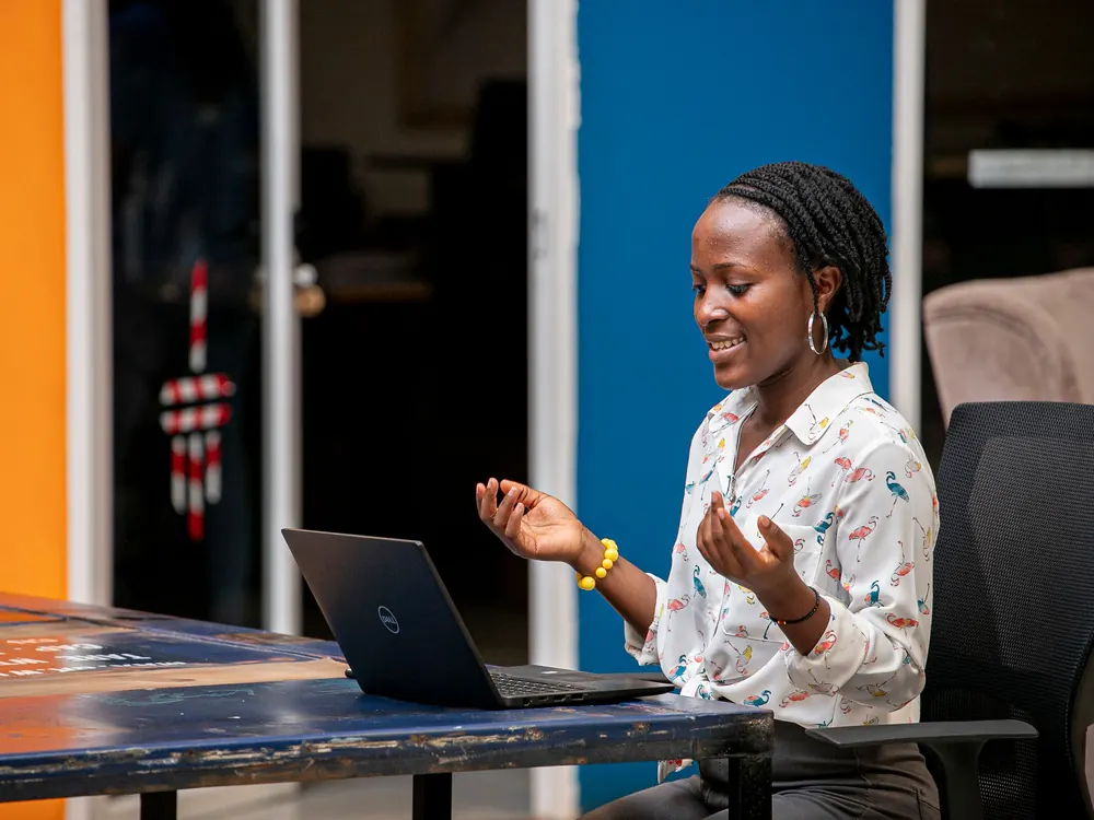 A young African intern speaking and gesturing to her laptop during a virtual team stand-up at a co-working desk