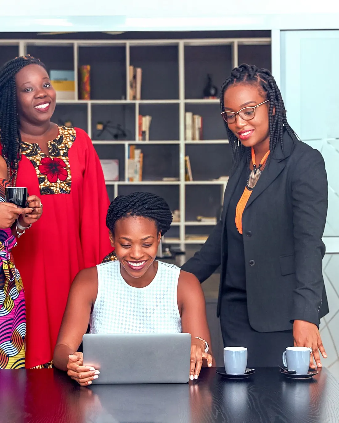 Three young African women working together around a laptop, one leaning in to guide the others